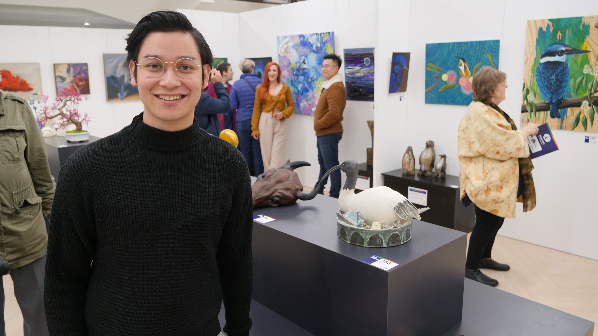 Matt at a ceramics exhibition, standing beside his ibis sculpture with paintings and other work behind him
